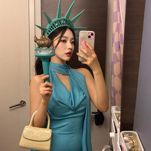 Woman wearing a teal sleeveless dress and Statue of Liberty crown holding a replica torch, posing for a selfie.