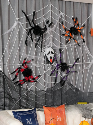 Halloween decorations featuring large colorful fuzzy spiders on a white spider web with a ghost face mask in the center on a gray background.